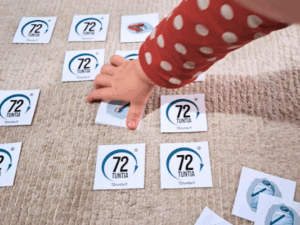 Muistipelikortteja, joissa 72 tuntia -logo) jaettuna alustalle kuvapuoli alaspäin. Pieni lapsen käsi tarttumassa yhteen korteista. Vieressä kuvapuoli ylöspäin saatu pari, jossa vesitonkan kuva.