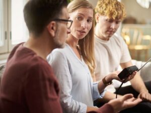 Nainen pitelee paristokäyttöistä radiota kädessään. Hänen vieressään istuvat nuori mies ja vanhempi mies.