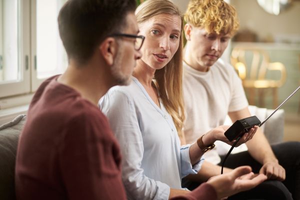 Nainen pitelee paristokäyttöistä radiota kädessään. Hänen vieressään istuvat nuori mies ja vanhempi mies.