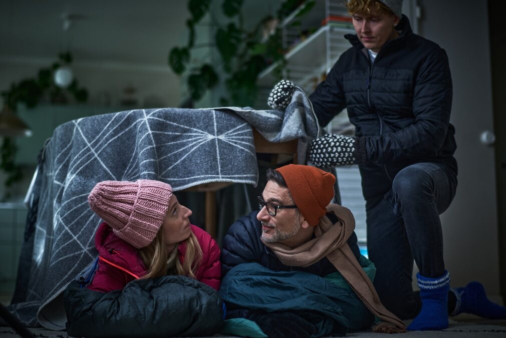 Sisällä ulkovaatteissa nainen ja mies vilteistä ja pöydästä tehdyn teltan sisällä. Ulkopuolella nuorempi poika myös ulkovaatteissa asettelee vilttiä pöydän päälle.