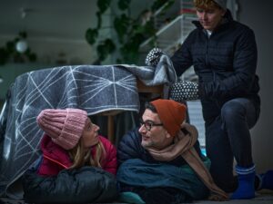 Sisällä ulkovaatteissa nainen ja mies vilteistä ja pöydästä tehdyn teltan sisällä. Ulkopuolella nuorempi poika myös ulkovaatteissa asettelee vilttiä pöydän päälle.